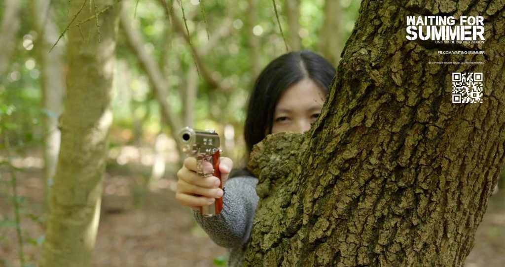 An image of a young girl hidden behind a trea, we only see her eyes and her right arm as she points her silver gun.